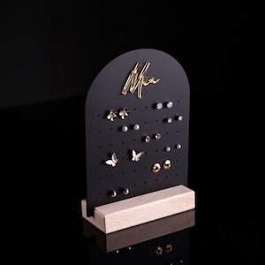 Op de afbeelding: Zwarte oorbellenstandaard met een houten basis. De standaard heeft een gebogen bovenkant met het woord "Mia" in gouden letters. De standaard is gevuld met verschillende oorbellen, waaronder vlinders, bloemen en studs.