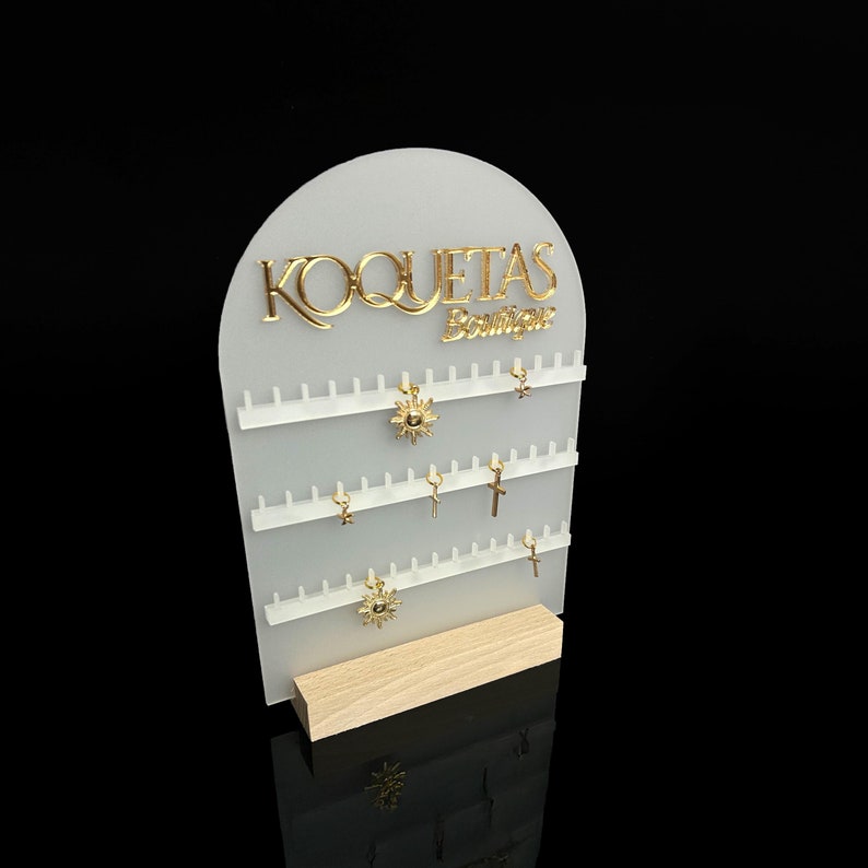 Puede incluir: Expositor de acr&iacute;lico blanco con el texto "KOQUETAS Boutique" en letras doradas. El expositor tiene varias filas de ganchos para exhibir joyas, con varios abalorios dorados colgando de los ganchos.