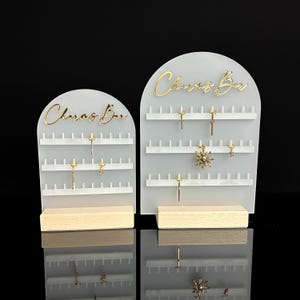 Puede incluir: Dos expositores de pendientes de acr&iacute;lico blanco con base de madera. Cada expositor tiene las palabras "Charms Bar" en letras doradas y varias filas de clavijas para colgar pendientes.