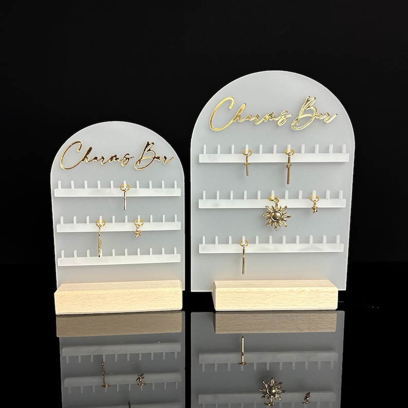 Puede incluir: Dos expositores de pendientes de acr&iacute;lico blanco con base de madera. Cada expositor tiene las palabras "Charms Bar" en letras doradas y varias filas de clavijas para colgar pendientes.