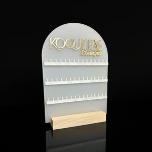 Puede incluir: Soporte de exhibici&oacute;n de aretes de acr&iacute;lico blanco con tres filas de clavijas. El soporte tiene una parte superior de arco redondeado con el texto "KOQUETAS Boutique" en letras doradas.