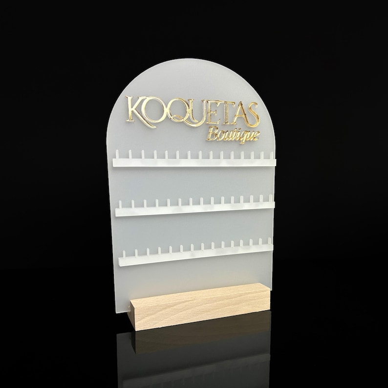 Puede incluir: Soporte de exhibici&oacute;n de aretes de acr&iacute;lico blanco con tres filas de clavijas. El soporte tiene una parte superior de arco redondeado con el texto "KOQUETAS Boutique" en letras doradas.