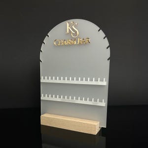 Puede incluir: Un expositor de joyas de acrílico transparente con el texto "KS CHARM BAR" en letras doradas. El expositor tiene varias filas de pequeñas clavijas para colgar joyas y una base de madera. El fondo es negro.