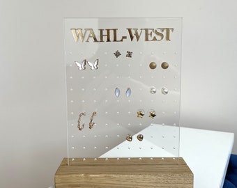 May include: A clear acrylic earring holder with a wooden base. The holder has a grid of holes for storing earrings. The text "WAHL-WEST" is printed in gold on the front of the holder.