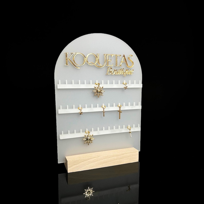 Puede incluir: Expositor de joyer&iacute;a de acr&iacute;lico blanco con el texto "KOQUETAS Boutique" en letras doradas. El expositor tiene varias filas de ganchos para exhibir pendientes. Hay varios pendientes dorados en el expositor, incluyendo dise&ntilde;os de sol y cruz.