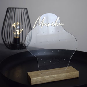 Op de afbeelding: Duidelijke acryl oorbellenhouder met een houten basis. De houder heeft een unieke vorm en heeft de naam "Marika" in zilveren letters.