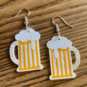 May include: A pair of novelty earrings shaped like beer mugs. The earrings feature a white mug with yellow beer and a white glittery foam top. The earrings have silver-colored fish hook ear wires.