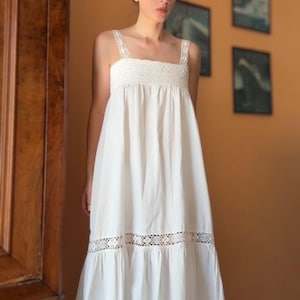 May include: A white midi-length dress featuring a square neckline, thick shoulder straps, and a smocked bodice. The garment includes a horizontal band of intricate floral lace detailing above the hemline, which finishes in a gathered ruffle.