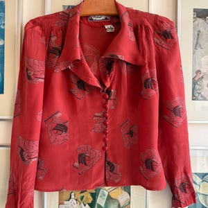 Peut inclure: Un chemisier rouge vintage avec un col et des manches longues. Le chemisier présente un motif répété de motifs de navires stylisés noirs et gris. Il a une fermeture boutonnée sur le devant et est suspendu à un cintre en bois.