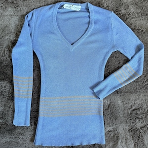 May include: A light blue long-sleeve sweater with a V-neck. The sweater has horizontal tan stripes on the lower torso and on the sleeves. The label reads "JACQUES HEIM PARIS".