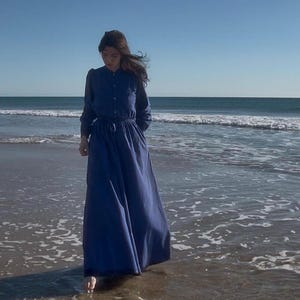 Peut inclure: Une longue robe bleu indigo fluide, à manches longues et boutonnage sur le devant. La robe est cintrée à la taille par une ceinture assortie. La personne se tient dans l'océan, les vagues se déferlant doucement sur ses pieds.
