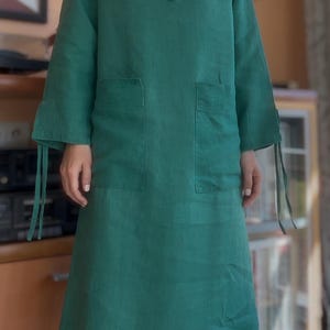 Puede incluir: Vestido de lino verde esmeralda con capucha, mangas largas y dos bolsillos delanteros. El vestido tiene cierres de lazo en el cuello y los puños. El vestido es hasta la rodilla y tiene un corte holgado.