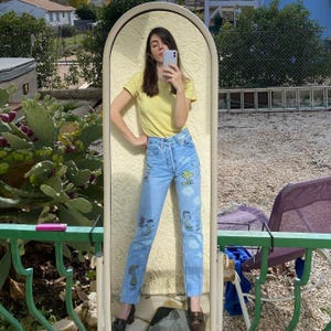 May include: Light blue denim jeans with cartoon character graphics. The jeans have a high waist and a button fly. The person is wearing a yellow t-shirt and black shoes. The mirror has an arched top.