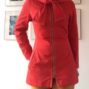 Peut inclure: Une veste rouge avec un grand nœud au col et une fermeture éclair sur le devant. La veste est ornée de coutures blanches sur les bords.