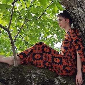 May include: A woman wearing a long, orange and black patterned dress with a geometric design. She is sitting on a tree branch with green leaves in the background.