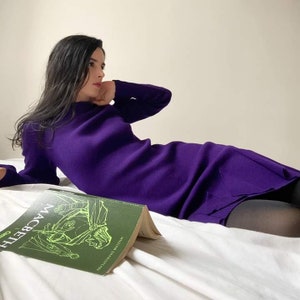 May include: A woman wearing a purple sweater dress is lying on a white bed with a green book titled "Macbeth" by William Shakespeare.