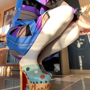 Peut inclure: Une femme portant une robe à motifs bleus, violets et bruns avec des collants blancs et des sabots plateforme bleus et blancs à talons rouges. Les sabots ont un motif à pois et le mot "Chopin" écrit sur le côté.