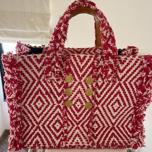 May include: A red and white woven tote bag with a geometric diamond pattern. The bag has a brown leather handle and gold-coloured circular embellishments. The edges are fringed, adding texture. A stylish accessory.