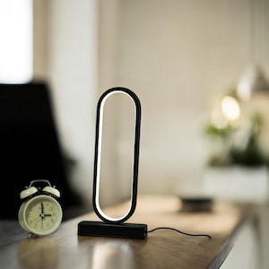 May include: A modern black LED desk lamp with a white illuminated oval design. The lamp is on a black base and has a power cord. A small, cream-colored alarm clock is to the left of the lamp.