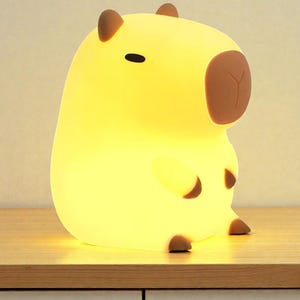 May include: A glowing yellow capybara-shaped night light. The lamp has brown accents on its ears, nose, and feet. The light is sitting on a wooden surface.
