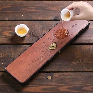May include: A wooden tea tray with a carved design, featuring a central floral motif and a decorative fan. A small white teacup sits beside the tray. Tea is being poured from a small cup onto the tray.
