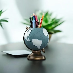May include: A globe-shaped desk organizer in shades of blue and white, holding various colored pencils and pens. The organizer sits on a bronze-colored base. A small white card and potted plants are in the background.