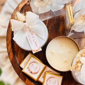 Może przedstawiać: Drewniana taca z różnymi upominkami ślubnymi. Upominki obejmują małe białe świece z perłowymi akcentami, małe białe pudełka z białą wstążką i suszonymi kwiatami oraz małe złote pudełka z kwiatowym wzorem i tekstem "Gift & Gather".