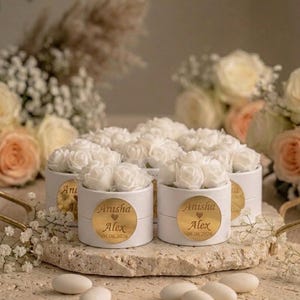Op de afbeelding: Witte, ronde dozen met witte rozen, geplaatst op een stenen oppervlak. Elke doos heeft een gouden sticker met de namen "Anisha" en "Alex" en de datum "06.06.2026". Verspreide snoepjes en bloemen op de achtergrond.