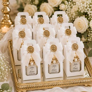 May include: A collection of white rectangular gift boxes with handles, each featuring a sheer white ribbon bow, a gold wax seal with an interlocking ring design, and a label printed with the text John, Liana, and the date 08.08.2026.