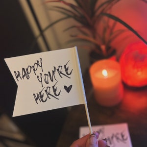 May include: A white flag with black lettering that reads "Happy you're here" with a small heart symbol. The flag is attached to a wooden stick and held by a hand.