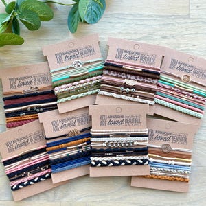 Puede incluir: Una colección de pulseras elásticas en varios colores, incluyendo negro, rosa, azul y marrón, exhibidas en tarjetas de cartón. Cada tarjeta tiene el texto "YOU ARE loved, BEAUTIFUL & ADORED". Algunas pulseras presentan cuentas y dijes decorativos.