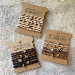 May include: Set of six brown and beige hair ties with gold and rhinestone accents. The hair ties are packaged on a card with the text "YOU ARE loved AWESOME wonderful BEAUTIFUL & ADORED".