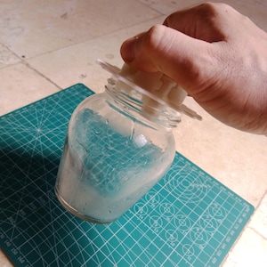 May include: A clear glass jar with a wide mouth and a threaded neck, featuring a white plastic lid attachment with a circular top and protruding tabs. The jar contains a light blue, semi-translucent substance with visible condensation on the interior glass.