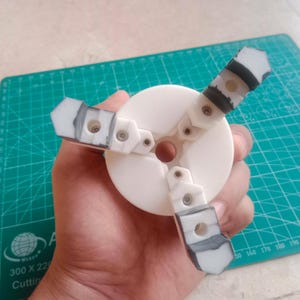 May include: A white circular mechanical component featuring three articulated arms extending from a central hub. Each arm is constructed from white segments connected by small metal fasteners, with dark gray textured tips at the ends of each arm.