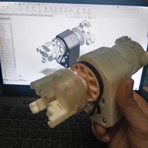Może przedstawiać: Wydrukowany w 3D zespół mechaniczny z białą plastikową obudową, centralną zębatką w kolorze brzoskwiniowym i czarnym gumowym paskiem. Urządzenie zawiera uchwyt szczękowy i jest trzymane przed ekranem komputera z modelem CAD.