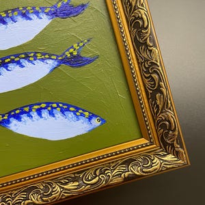May include: An oil painting of three stylized fish with blue and yellow markings on a green background. The fish are set in a gold ornate frame. The painting has a textured surface.