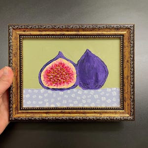 Könnte beinhalten: Gerahmtes Gemälde mit zwei Feigen. Eine Feige ist aufgeschnitten und zeigt ein rosa-gelbes Inneres. Die andere Feige ist ganz. Der Hintergrund ist hellgrün, der Tisch weiß mit blauem Muster. Der Rahmen ist goldfarben.