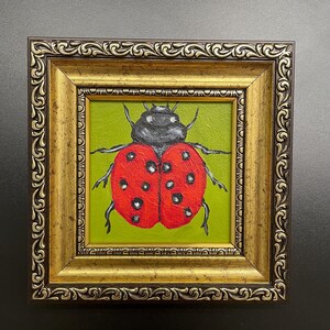 May include: A framed painting of a ladybug. The ladybug has a red shell with black spots and a black head. The background is green. The frame is gold with ornate detailing.