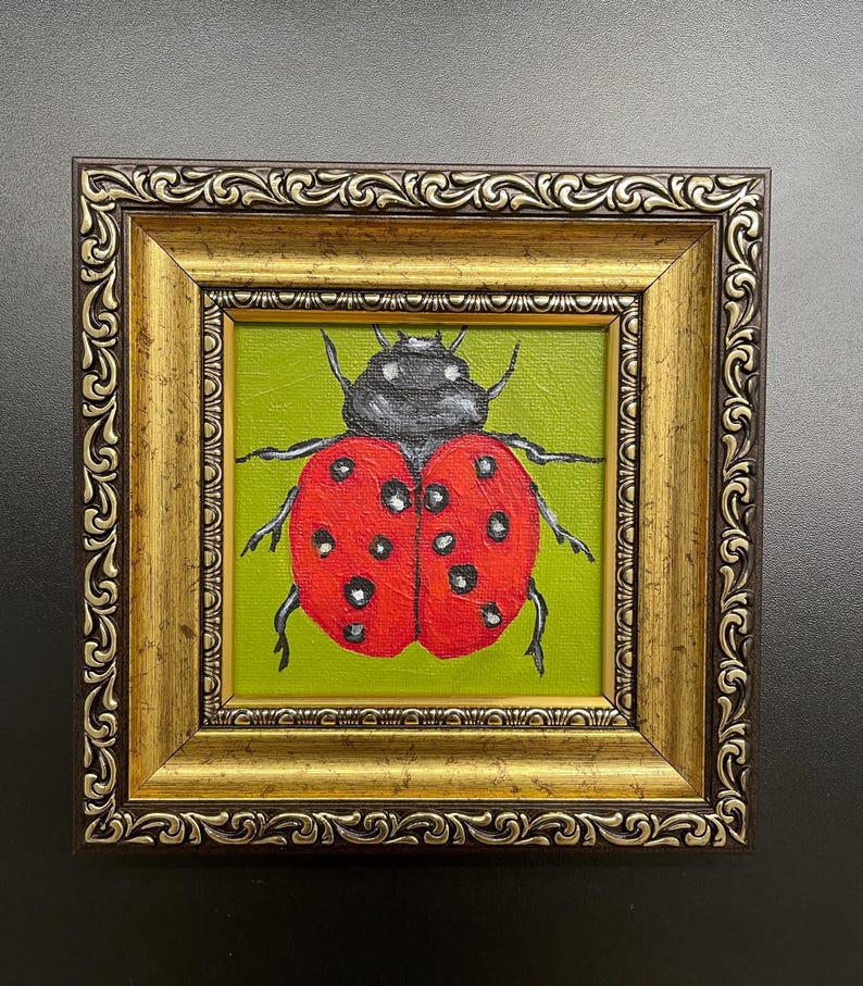 May include: A framed painting of a ladybug. The ladybug has a red shell with black spots and a black head. The background is green. The frame is gold with ornate detailing.