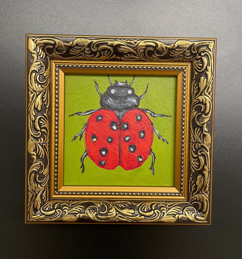 May include: A framed painting of a ladybug. The ladybug is red with black spots and a black head, set against a green background. The ornate frame is gold and dark brown.