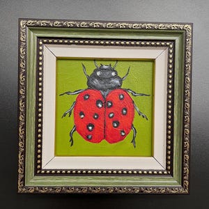 May include: A framed painting of a ladybug. The ladybug is red with black spots and a black head, set against a green background. The frame is green with gold accents and a white inner border.
