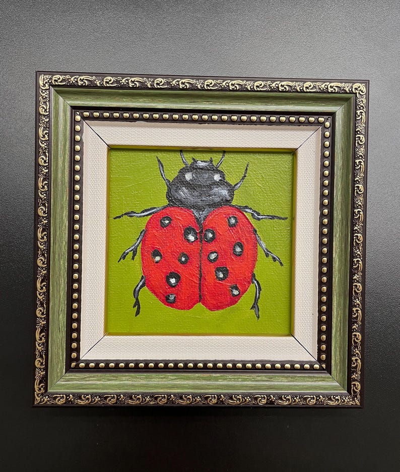May include: A framed painting of a ladybug. The ladybug is red with black spots and a black head, set against a green background. The frame is green with gold accents and a white inner border.