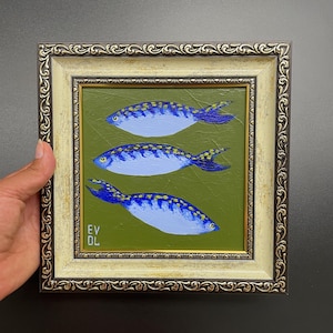 May include: A framed painting featuring three blue fish with yellow spots on a green background. The fish are painted with white highlights and dark blue tails. The frame is ornate with a gold and cream color.