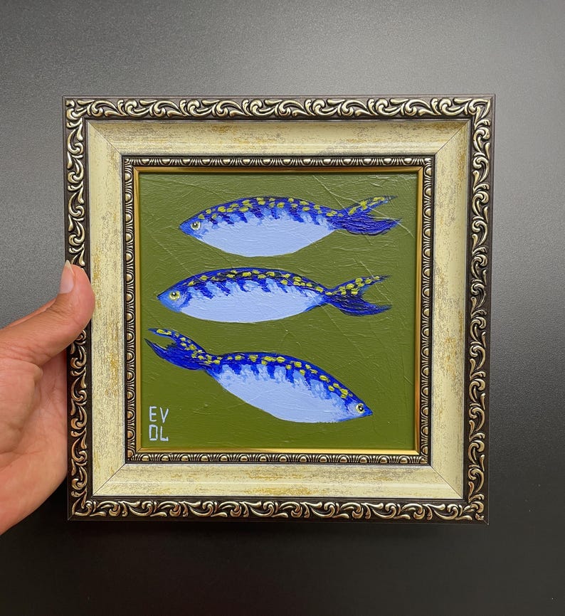 May include: A framed painting featuring three blue fish with yellow spots on a green background. The fish are painted with white highlights and dark blue tails. The frame is ornate with a gold and cream color.