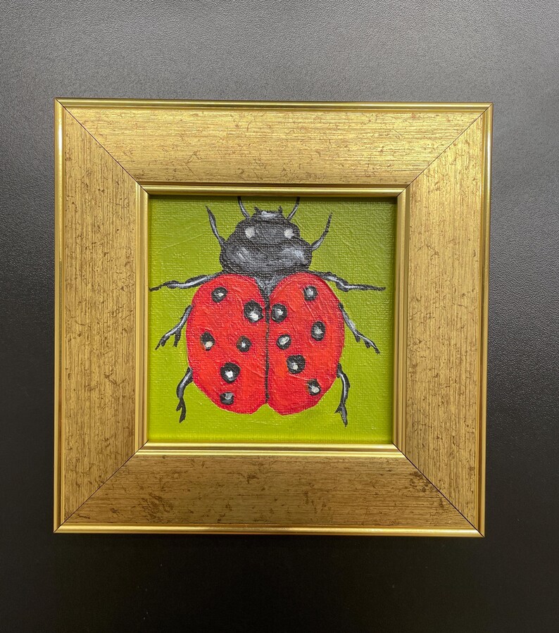 May include: A framed painting of a ladybug. The ladybug is red with black spots and a black head, set against a green background. The painting is in a gold-colored frame, creating a decorative piece.