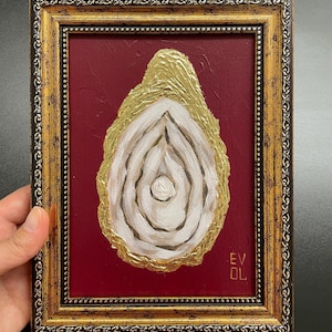 May include: An abstract painting in a gold ornate frame. The artwork features a white and gold oyster-like shape on a red background. The frame is gold with a decorative pattern. The artist's initials are in the bottom right corner.