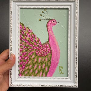 May include: A framed painting of a pink peacock with green and pink feathers against a light green background. The peacock is in a white ornate frame. The painting has the initials "EV DL" in the bottom right corner.