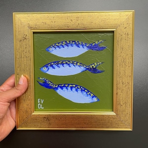 May include: A framed painting featuring three blue fish with yellow spots on a green background. The fish have dark blue fins and tails. The frame is gold-colored and rectangular. The artist's initials, "EV DL", are in the lower left corner.