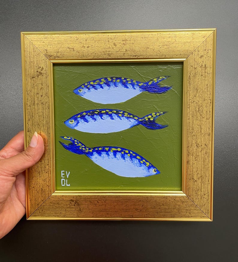 May include: A framed painting featuring three blue fish with yellow spots on a green background. The fish have dark blue fins and tails. The frame is gold-colored and rectangular. The artist's initials, "EV DL", are in the lower left corner.