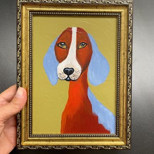 May include: A framed painting of a dog with a red and white face, blue ears, and a red neck. The dog is set against a yellow-green background. The frame is gold with ornate detailing.